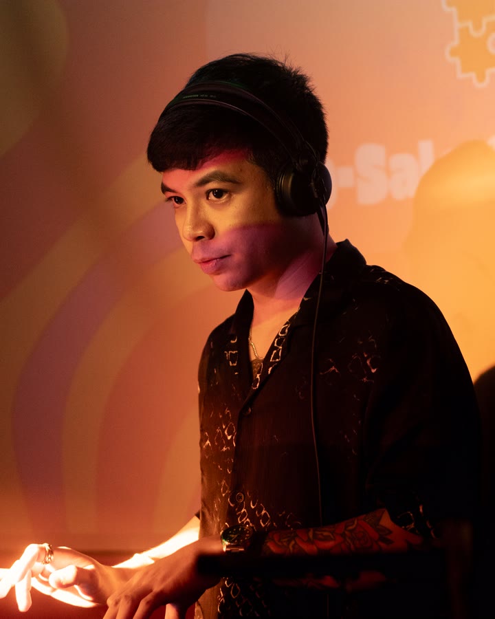 REIDEL at the decks, headphones on, looking up from the controller under warm light.